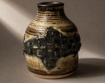 May include: A ceramic vase with a rounded body and a narrow neck, featuring a beige and brown speckled glaze. A prominent, textured band of dark blue, square-shaped indentations wraps around the center of the vessel, creating a contrast.