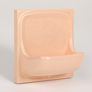 Może przedstawiać: Brzoskwiniowa mydelniczka ścienna. Mydelniczka ma kwadratowy tył z zakrzywioną, prostokątną misą i zakrzywioną przednią krawędzią. Mydelniczka jest wykonana z ceramiki i ma błyszczące wykończenie.