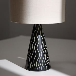 May include: A table lamp featuring a conical black base decorated with vertical, wavy white lines. The lamp is topped with a cylindrical, off-white fabric lampshade that provides a neutral contrast to the dark, patterned base below it.
