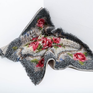 Peut inclure: Un tapis gris et blanc avec un motif floral de roses rouges et de feuilles vertes. Le tapis a une texture touffue et une bordure à franges. Le tapis est plié dans un coin.