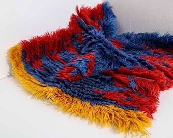 May include: A fluffy rug with a geometric pattern in shades of red, blue, and yellow. The rug has a fringe border.