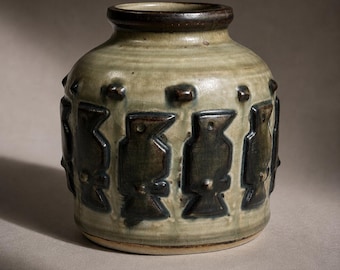 Peut inclure: Un vase en céramique avec un corps arrondi et un bord marron foncé. Le vase est vert clair avec des figures sombres en forme d'oiseaux autour du corps. De petits points en relief sont placés au-dessus des figures. La base est d'une couleur plus claire.