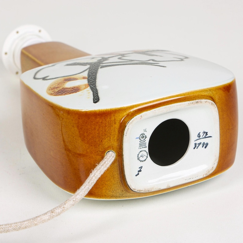 May include: A brown and white ceramic table lamp with a black floral design. The lamp has a cord and a square base with a round hole in the center. The base is marked with the numbers "6 73 37 99".