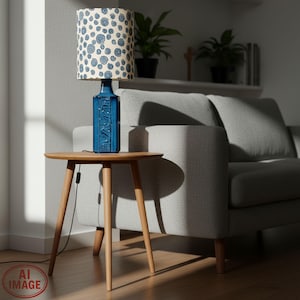 Puede incluir: Una lámpara de mesa de cerámica azul con una pantalla crema con un patrón de espirales azules. La lámpara está sobre una pequeña mesa auxiliar redonda de madera con tres patas. La base de la lámpara tiene un diseño texturizado. Un sofá gris está cerca.