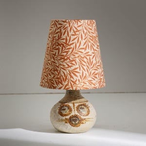 Puede incluir: Una pequeña lámpara de mesa con base de cerámica beige y pantalla cónica. La pantalla presenta un patrón repetido de hojas naranjas sobre fondo crema. La base tiene forma redondeada con diseños circulares marrones.