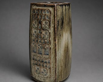 Peut inclure: Un vase en céramique rectangulaire avec une glaçure marbrée vert olive et marron. Le panneau avant présente une grille de carrés en creux, chacun contenant un petit motif floral ou géométrique en relief dans des tons bleu foncé et marron.