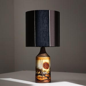 May include: A table lamp featuring a cylindrical ceramic base with a hand-painted landscape scene in shades of brown, tan, and black, topped with a dark brown drum lampshade that has vertical textured panels and thin light-colored accent lines.