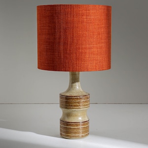 Puede incluir: Una lámpara de mesa de cerámica con una pantalla naranja cilíndrica. La base es beige con rayas horizontales marrones. La lámpara tiene un diseño moderno de mediados de siglo.