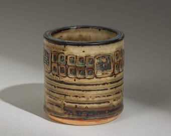 Peut inclure: Une tasse cylindrique en céramique aux tons beige et marron. La tasse présente une série de motifs rectangulaires au milieu, avec des lignes horizontales et un bord foncé. La base est de couleur brun rougeâtre.