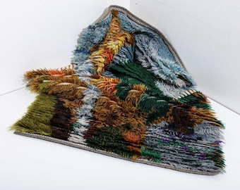 May include: A colorful, textured rug with a variety of colors including blue, green, brown, orange, and purple. The rug has a shaggy, fringed appearance and a gray border. The rug is folded over on itself, showing the different colors and textures.