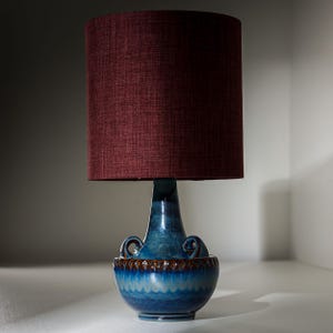 Puede incluir: Una lámpara de mesa vintage con una pantalla de tela burdeos. La base es de cerámica azul vibrante con un acabado texturizado y vidriado y asas decorativas. La lámpara tiene un diseño retro único.