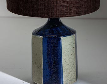 Peut inclure: Une lampe de table en céramique avec un abat-jour marron foncé. Le pied de la lampe est de forme géométrique avec des rayures bleues et blanc cassé alternées. La lampe est conçue pour une utilisation en intérieur.