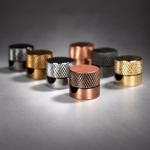 Peut inclure: Un ensemble de sept boutons cylindriques en métal disposés en groupe. Chaque bouton présente une partie supérieure texturée avec un motif en losange et une base lisse et solide avec une petite découpe semi-circulaire. Les finitions incluent des tons cuivre, laiton, argent et gris foncé.