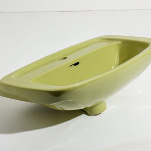 May include: A pale green ceramic bathroom sink with a rectangular basin and rounded edges. The sink features a built-in tap hole and drain, and rests on a small pedestal. The design is sleek and modern.
