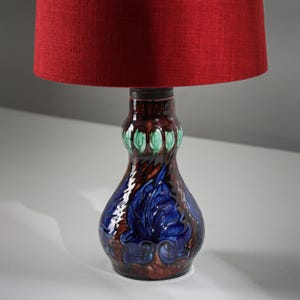 Puede incluir: Lámpara de mesa decorativa con pantalla roja. La base de cerámica presenta un diseño floral marrón, azul y verde. La lámpara tiene una estética clásica y vintage, adecuada para una sala de estar o dormitorio.