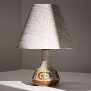 May include: A table lamp featuring a textured, off-white ceramic base with a wide, rounded bottom and a tapered neck. The base is decorated with a repeating brown circular pattern. It is topped with a conical, light gray fabric lampshade.