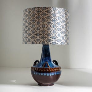 Puede incluir: Una lámpara de mesa de cerámica con base azul y marrón y una pantalla con estampado geométrico. La pantalla presenta un patrón repetitivo de formas azules en forma de estrella sobre un fondo crema. La lámpara tiene dos asas pequeñas a los lados.