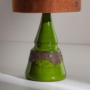 Puede incluir: Una lámpara de mesa de cerámica verde con una banda texturizada marrón alrededor de la base y una pantalla de color óxido. La lámpara tiene un acabado brillante y un diseño artístico único, adecuado para la decoración del hogar.