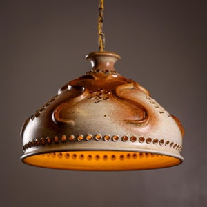 Puede incluir: Una lámpara colgante de cerámica con un esmalte marrón y blanco. La luz tiene una forma orgánica única con una superficie texturizada y una fila de agujeros alrededor del borde inferior. La luz está iluminada, proyectando un brillo cálido.