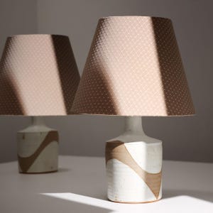 May include: Two table lamps featuring cylindrical ceramic bases with a white speckled glaze and a brown wave pattern. Each lamp is topped with a tapered, light brown fabric lampshade that has a subtle, repeating white diamond grid pattern.