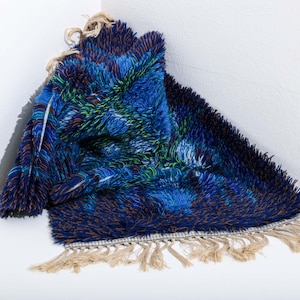 May include: A textured, abstract rug in shades of blue, green, and brown. The rug has a shaggy appearance with a fringe along the edge. The rug is folded in a corner, showcasing its design and texture.