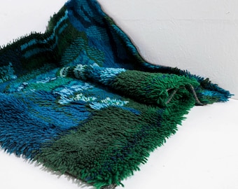 May include: A blue and green shag rug with a geometric pattern. The rug is folded in half and is lying on a white surface.