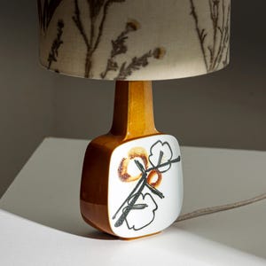 May include: A ceramic table lamp with a brown and white base and a floral patterned lampshade. The base features a stylized floral design in black and brown.