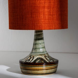 Puede incluir: Una lámpara de mesa de cerámica con una pantalla de tela naranja quemado. La base es una mezcla de colores verde, marrón y crema con un patrón decorativo. La lámpara es una lámpara decorativa.