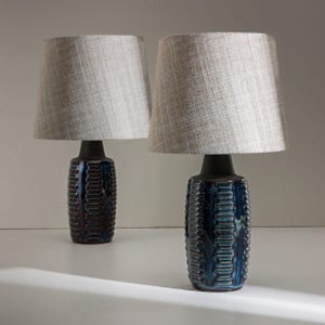 Puede incluir: Dos lámparas de mesa a juego. Cada lámpara tiene una base de cerámica azul oscuro texturizada con un cuello negro, rematada con una pantalla de tela gris claro. Las lámparas están diseñadas para uso en interiores y proporcionan iluminación ambiental.