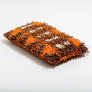 Peut inclure: Coussin décoratif rectangulaire avec une surface touffue et texturée. Le coussin présente un motif de zigzags orange, marron et blanc cassé. La base du coussin est de couleur orange unie.