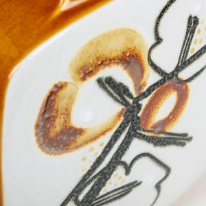 May include: Close-up of a ceramic dish with a brown glaze and a stylized floral design in black and brown. The design features a stylized flower with a curved stem and leaves.