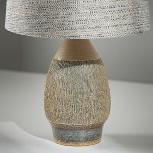 Puede incluir: Una lámpara de mesa de cerámica con una base texturizada beige y azul y una pantalla con estampado azul claro y blanco. La lámpara tiene un cuerpo redondeado y texturizado y un cuello de tono neutro.