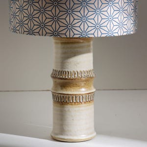 Puede incluir: Una lámpara de mesa de cerámica con base color crema y pantalla con estampado geométrico azul y blanco. La base tiene un diseño texturizado similar al bambú con detalles marrones. Lámpara decorativa.