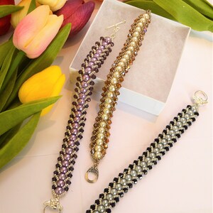 May include: Three beaded bracelets in shades of purple, gold, and silver with decorative clasps. The bracelets are displayed on a white surface with a box and colourful tulips in the background. The bracelets are approximately 18 cm long.