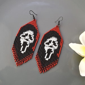 Op de afbeelding: Een paar kralen oorbellen in zwart, rood en wit. De oorbellen hebben een zwarte basis met een witte figuur in het midden. De randen zijn rood en de haken zijn zwart. Een uniek accessoire.