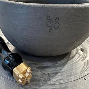 May include: A dark gray ceramic bowl with an embossed design and a brass stamp tool with a wooden handle. The bowl has a simple, rounded shape and a matte finish. The stamp tool is positioned near the bowl.