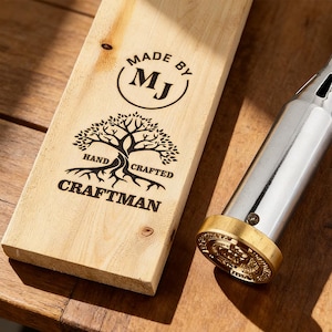 Op de afbeelding: Een houten plank met de tekst "MADE BY MJ HAND CRAFTED CRAFTMAN" en een boomlogo staat naast een metalen brandijzer met een messing kop. Het brandijzer is ontworpen voor houtbranden.