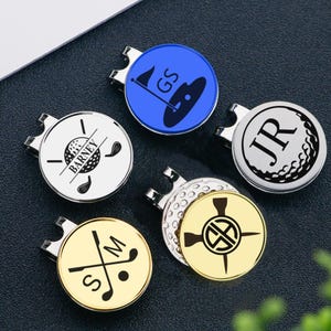 Könnte beinhalten: Eine Sammlung von Golfballmarkern mit Magnetclips. Die Marker zeigen verschiedene Designs, darunter einen Golfball, gekreuzte Schläger und Initialen. Farben sind Silber, Gold und Blau. Die Marker sind für den Einsatz auf dem Golfplatz konzipiert.