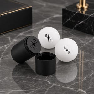 May include: Two white spherical balls and a black cylindrical container with a matching lid. Each ball features a black logo with the letters B and K separated by a four-pointed star. The lid also displays the same black logo design.