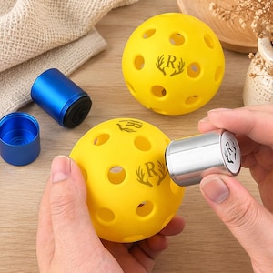 Könnte beinhalten: Zwei leuchtend gelbe Pickleballs mit mehreren kreisförmigen Löchern und einem aufgedruckten Logo, das den Buchstaben R zwischen zwei Geweihstangen zeigt. Ein zylindrischer Stempel aus silbernem Metall mit gleichem Logo markiert den Ball.