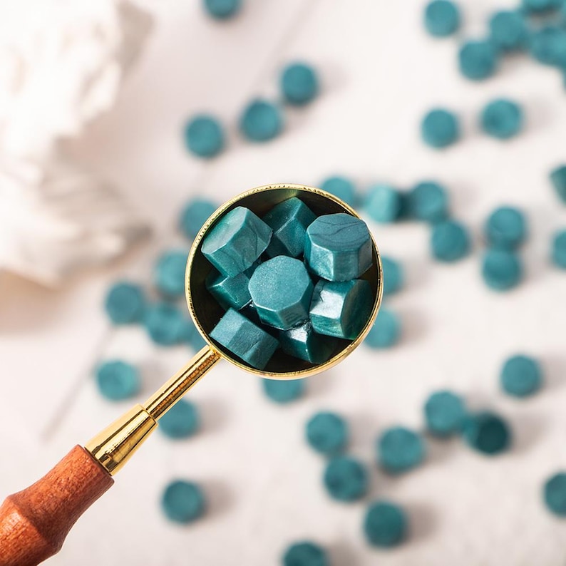May include: A gold metal spoon filled with teal blue wax sealing beads. The beads are hexagonal in shape and are scattered on a white surface.