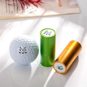 May include: Three golf ball markers, one white golf ball, and two small containers. The containers are green and gold. The white golf ball has a black and white golf club logo with the letters "RG". The green container has a white lid with a black and white golf club logo with the letters "RG". The gold container has a white lid with a green golf ball logo and the word "ALISA".