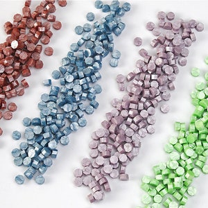 May include: Assortment of sealing wax beads in four colours: red, blue, purple, and green. The beads are arranged in rows on a white surface.