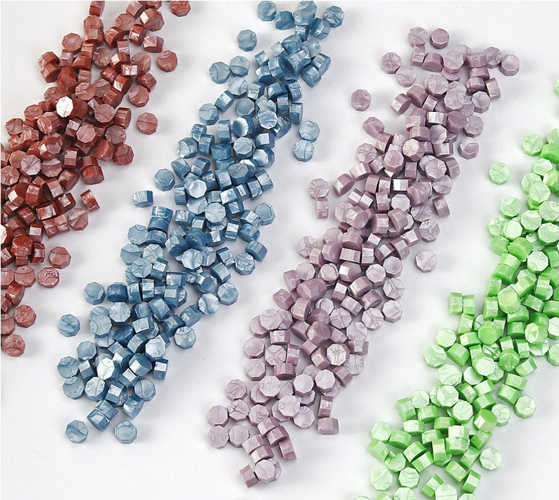 May include: Assortment of sealing wax beads in four colours: red, blue, purple, and green. The beads are arranged in rows on a white surface.