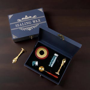 Può includere: Un set blu navy per ceralacca, con dettagli dorati e la scritta "SEALING WAX" sul coperchio. Il set include uno scaldacera, perline di cera, un cucchiaio e due strumenti decorativi dorati.