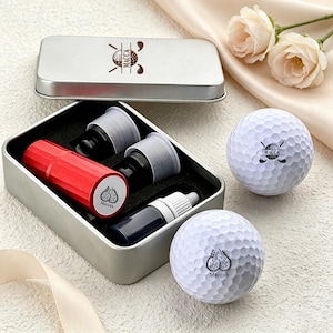May include: A silver metal rectangular tin containing a red cylindrical stamp, two black ink cartridges, and a small bottle of ink. Two white golf balls are placed nearby, each featuring a black graphic logo with the text Macca's printed on them.