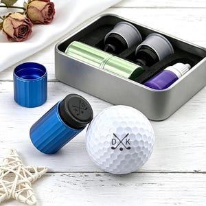 Puede incluir: Una pelota de golf blanca con las iniciales "DK" y palos de golf cruzados está junto a un marcador de pelota de golf azul y un soporte para marcador azul. Una caja de metal contiene un marcador verde, dos marcadores grises y una botella morada.