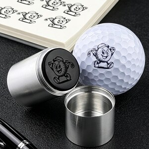 Könnte beinhalten: Ein silberner Golfball-Stempel aus Metall mit einem schwarzen, runden Stempelkopf, der eine Golfball-Cartoonfigur zeigt. Daneben liegt ein weißer Golfball mit Grübchen, auf dessen Oberfläche dieselbe Cartoonfigur schwarz aufgedruckt ist.