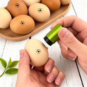 May include: An assortment of brown and beige eggs, some stamped with "Mindy's Eggs" and a chicken graphic. A green and black egg stamper is being used on an egg. The eggs are in a wooden tray, with a sprig of green leaves in the foreground.