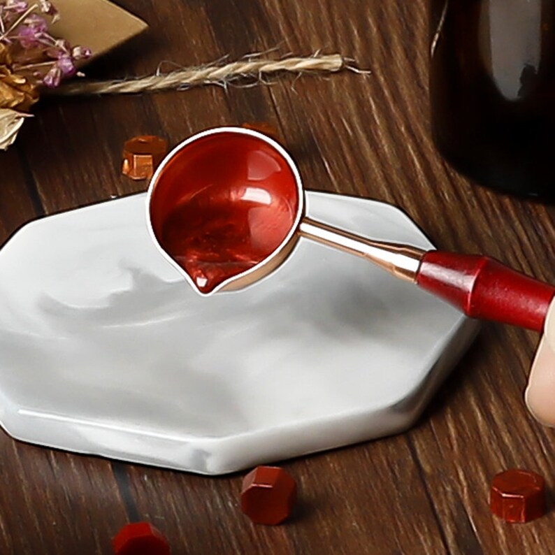May include: A red metal spoon with a wooden handle, used for melting wax seals. The spoon is resting on a white marble surface with red wax seal pellets nearby.